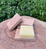 Handcrafted soap rests on a wooden soap dish, displayed on a textured tree stump outdoors.
