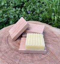 Handcrafted soap rests on a wooden soap dish, displayed on a textured tree stump outdoors.
