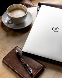 A Dell laptop sits on a wooden desk next to a cup of coffee, a leather notebook, and a pen, creating a workspace aesthetic.
