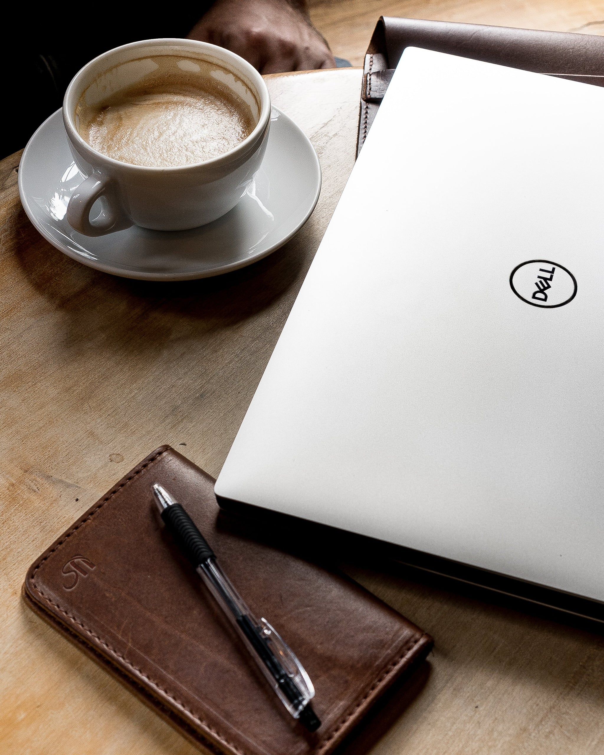 A Dell laptop sits on a wooden desk next to a cup of coffee, a leather notebook, and a pen, creating a workspace aesthetic.
