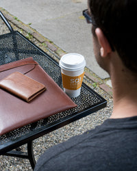 Man enjoys SQ1 Coffee Roasters coffee outdoors alongside his leather wallet and laptop case on a wrought iron table.
