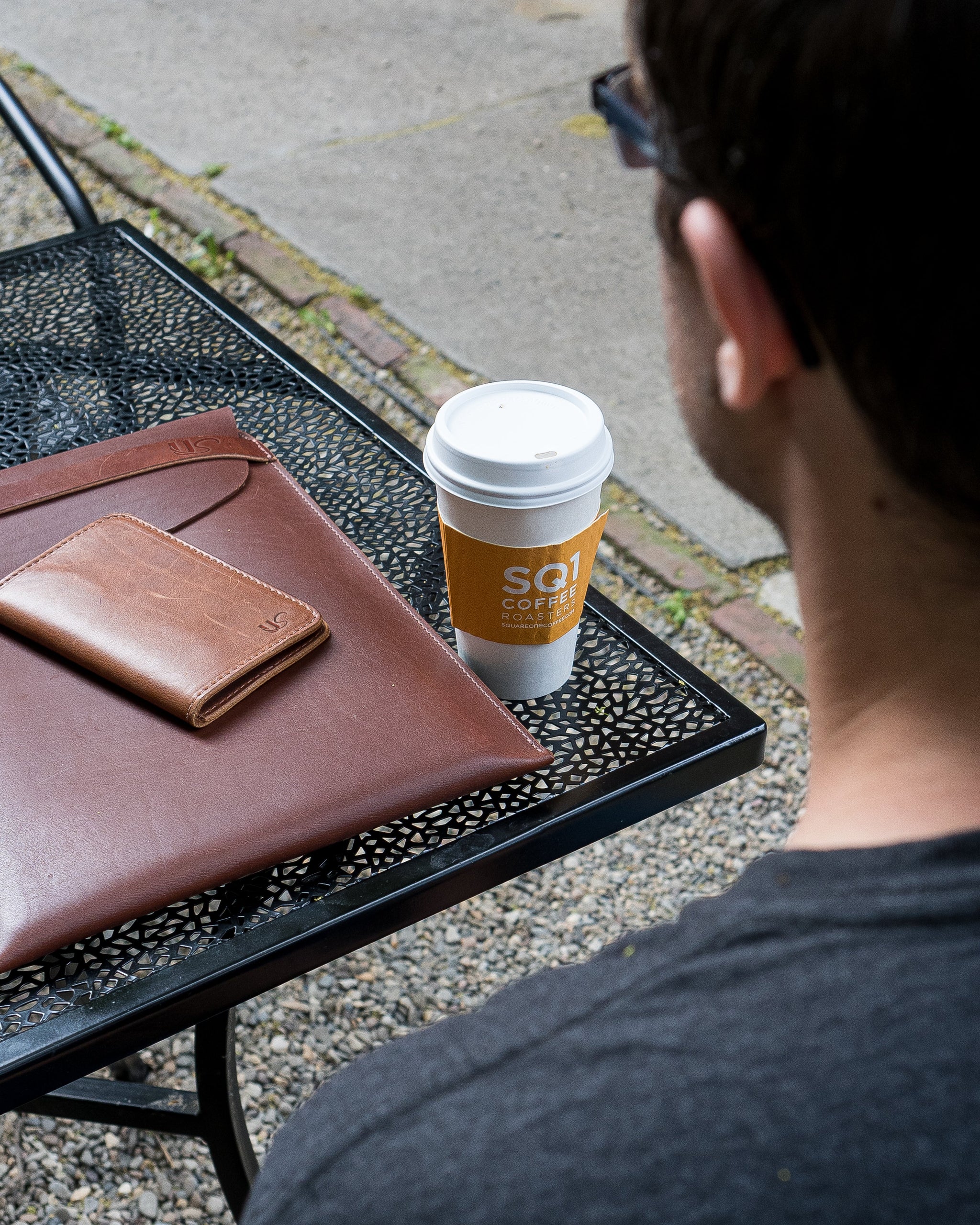 Man enjoys SQ1 Coffee Roasters coffee outdoors alongside his leather wallet and laptop case on a wrought iron table.
