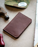 A handcrafted brown leather wallet sits on a workbench near a hammer, saw, and camouflage hat, suggesting artisan craftsmanship.
