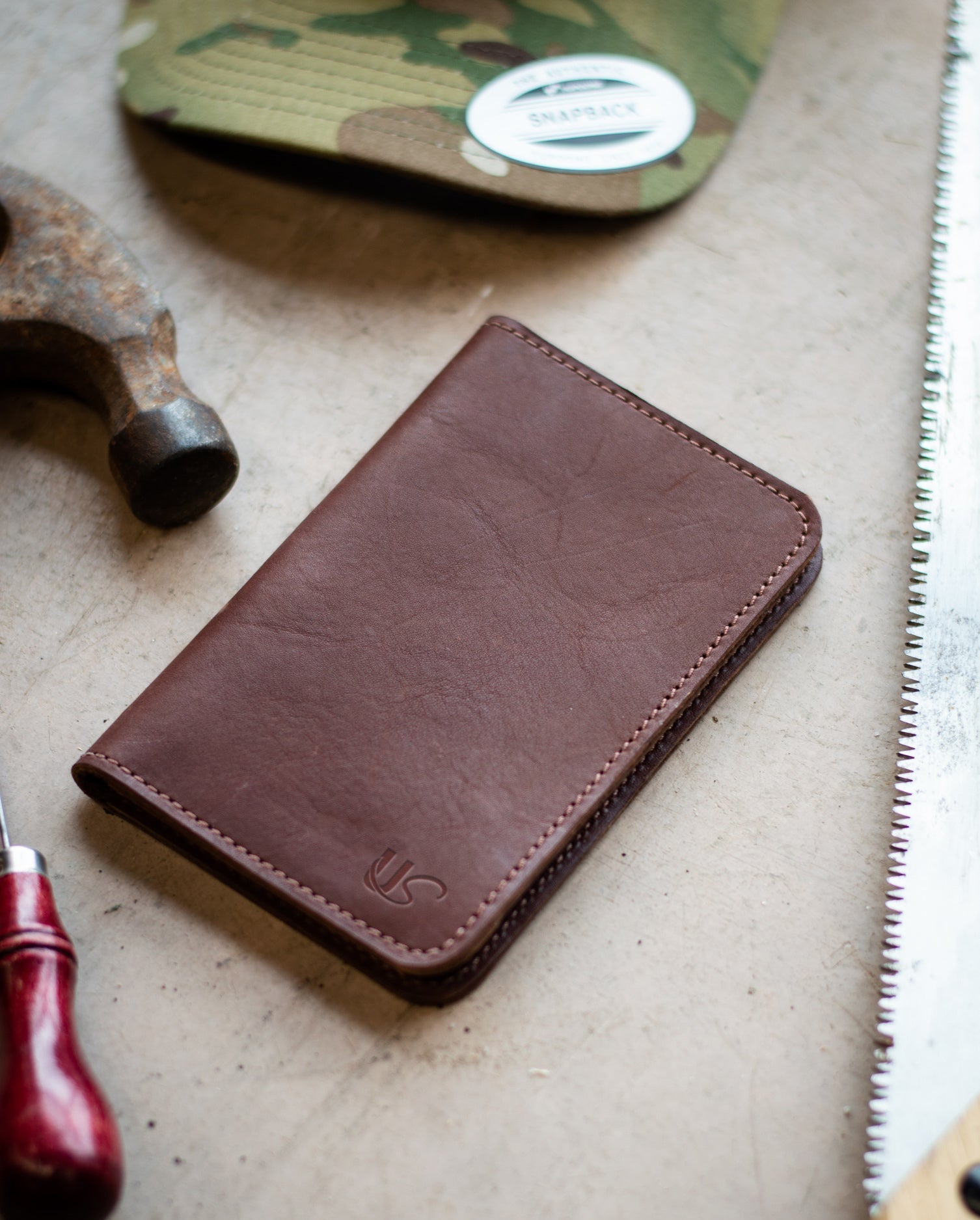 A handcrafted brown leather wallet sits on a workbench near a hammer, saw, and camouflage hat, suggesting artisan craftsmanship.
