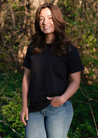 A brunette woman smiles outdoors, wearing a black t-shirt and jeans, with her hand casually in her pocket.
