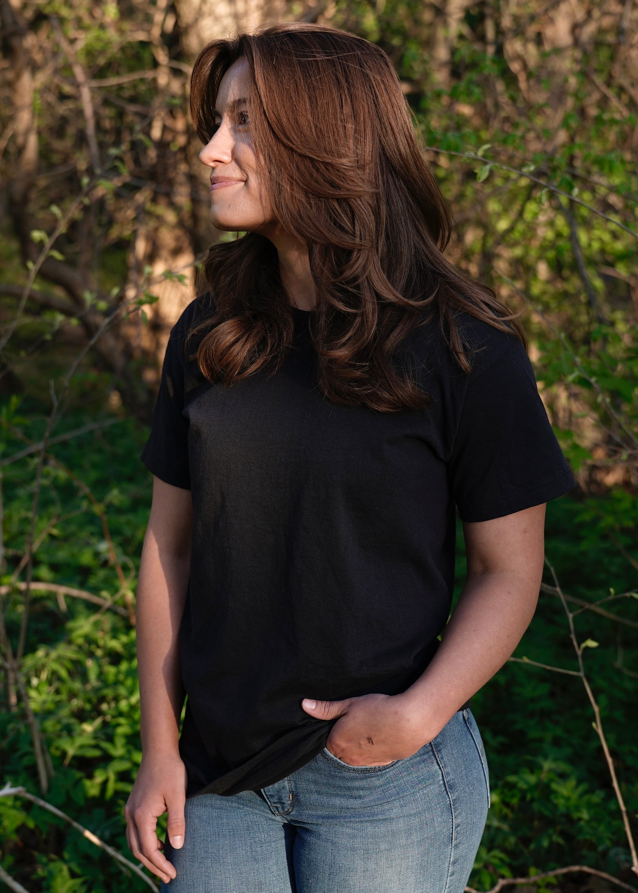 Woman wearing a black T-shirt and jeans looks thoughtfully toward sunlit foliage.