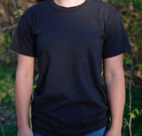A model wears a plain black T-shirt outdoors against a blurred green backdrop.
