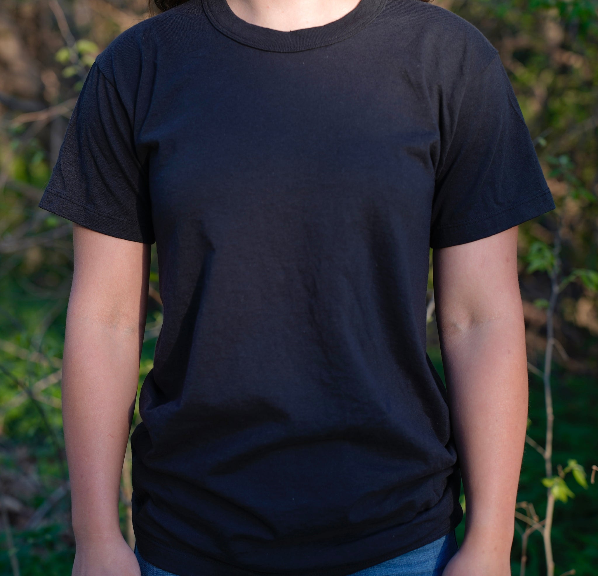 A model wears a plain black T-shirt outdoors against a blurred green backdrop.
