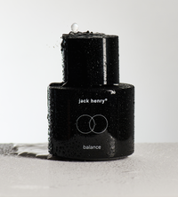 Water splashes onto a black Jack Henry Balance skincare product bottle.
