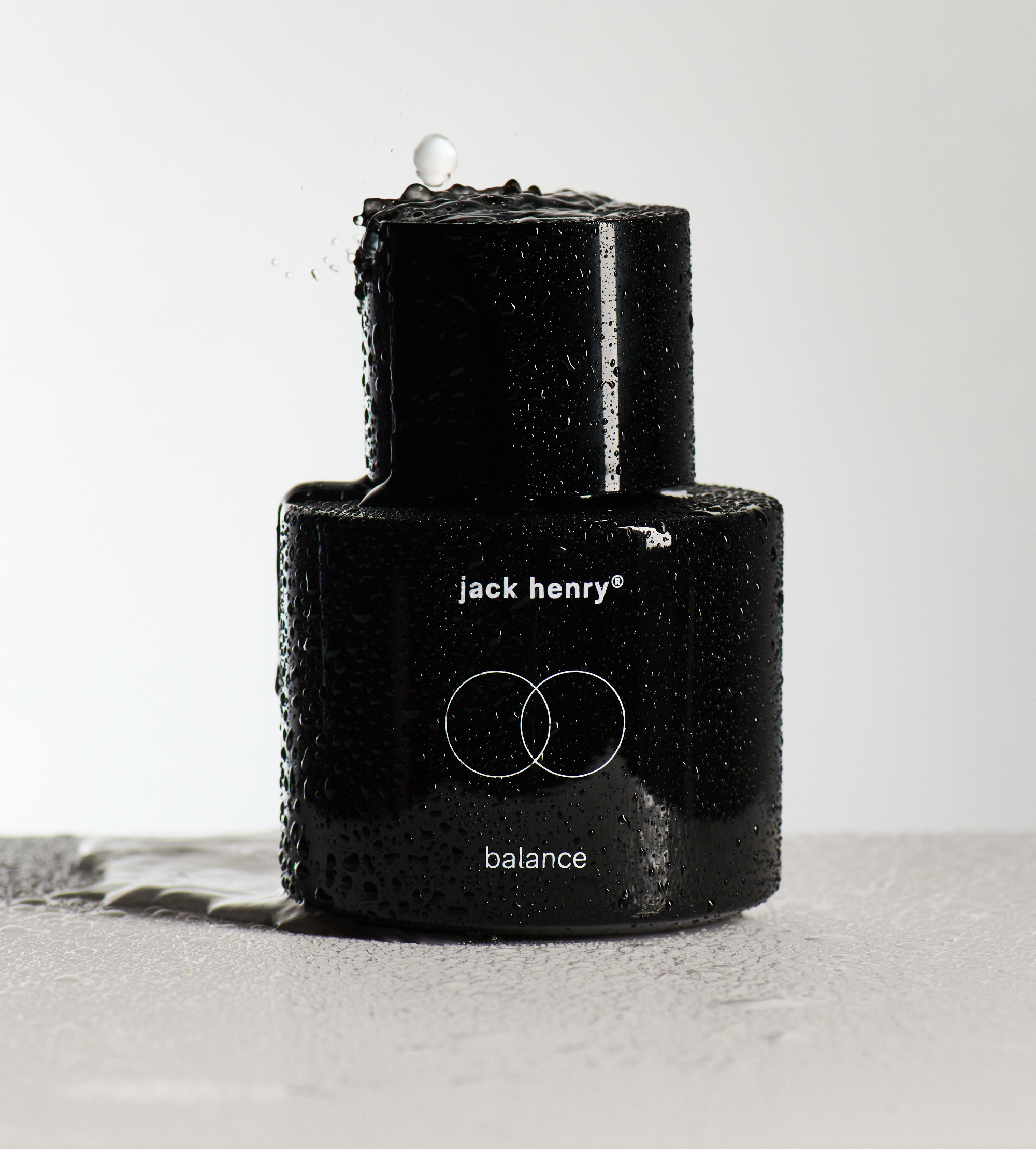 Water splashes onto a black Jack Henry Balance skincare product bottle.

