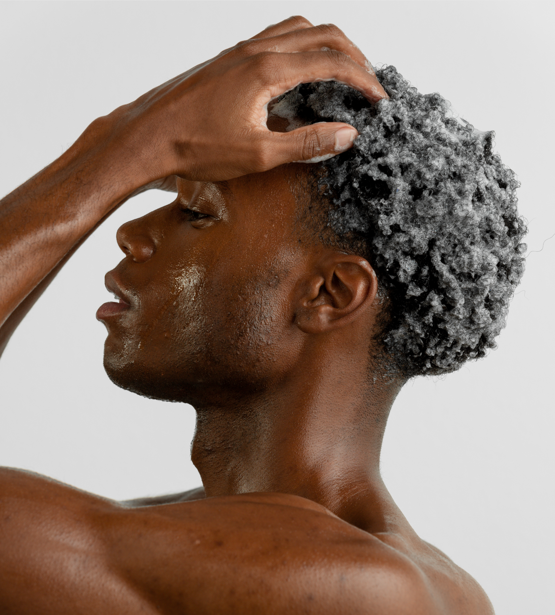 An African American man massages shampoo into his curly hair, showcasing his healthy skin.
