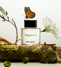 A monarch butterfly perches on a bottle of Jack Henry Frequency cologne displayed amid magnolia blossoms and mossy wood.
