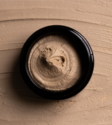 Creamy facial mud mask swirls inside a black container against a smeared earthy-toned background.
