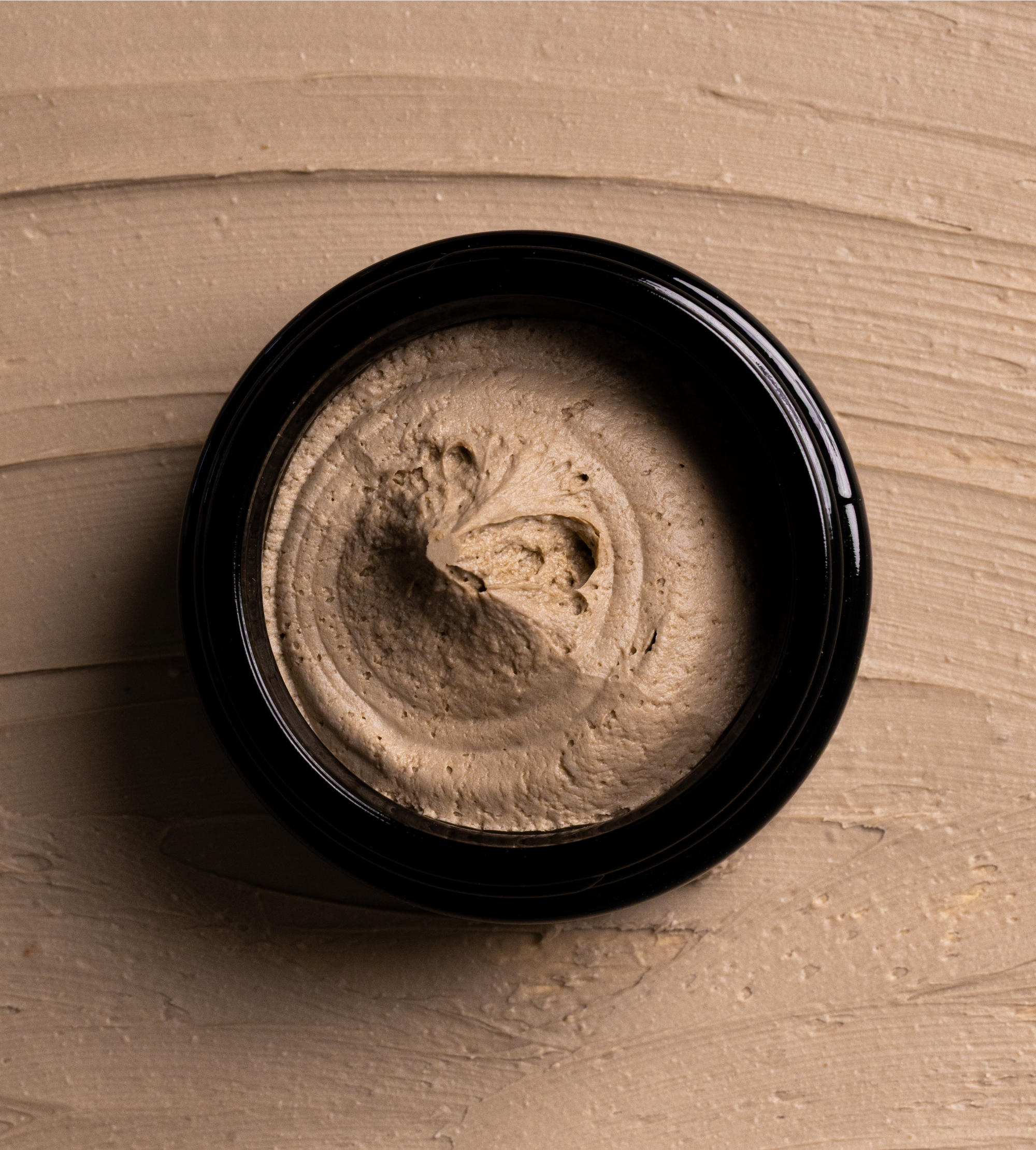 Creamy facial mud mask swirls inside a black container against a smeared earthy-toned background.
