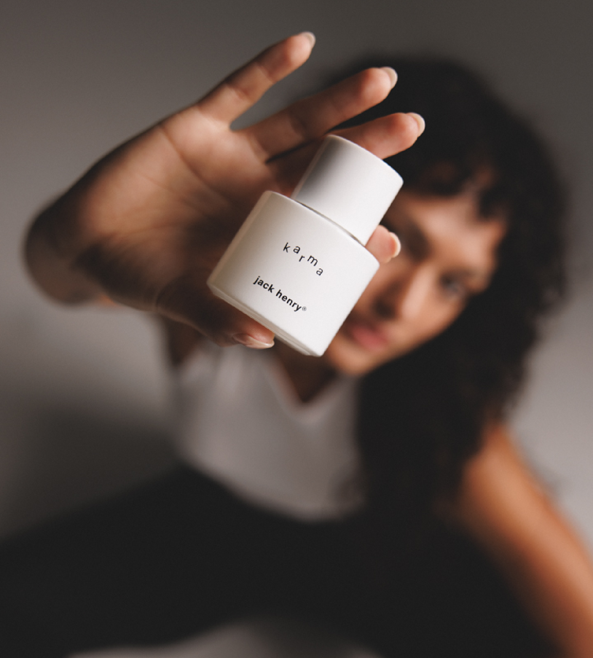 A woman presents a white "Karma" by Jack Henry® fragrance bottle toward the viewer.
