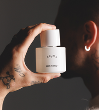 A tattooed hand holds a bottle of Jack Henry Karma fragrance up to a person's nose.
