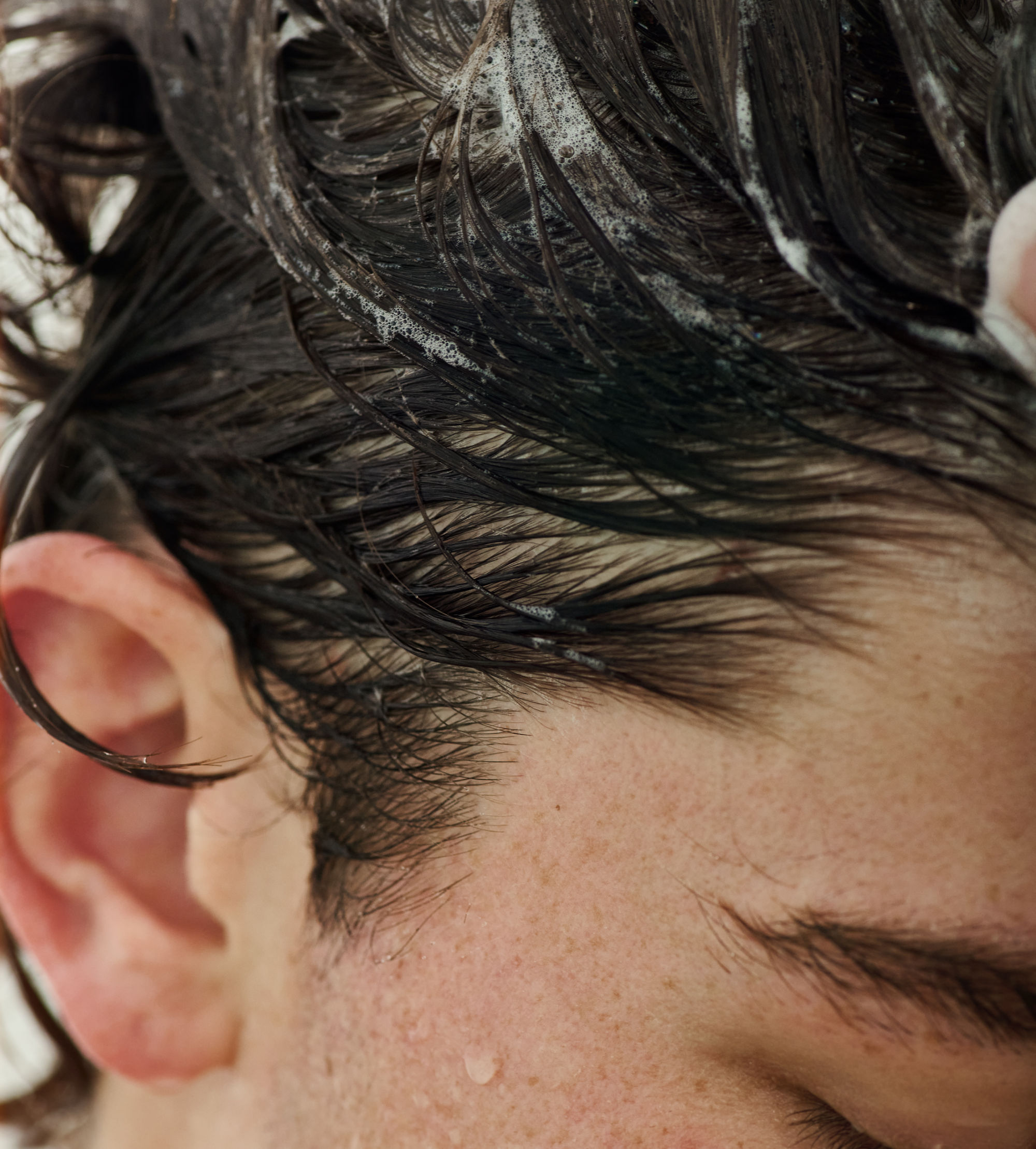 Close-up shows someone shampooing dark hair with foaming cleanser.
