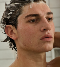 A young man washes his hair with shampoo in the shower, looking thoughtfully ahead.