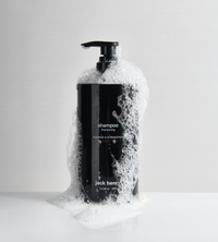 Jack Henry shampoo bottle overflows with cleansing bubbles on a glossy white surface.
