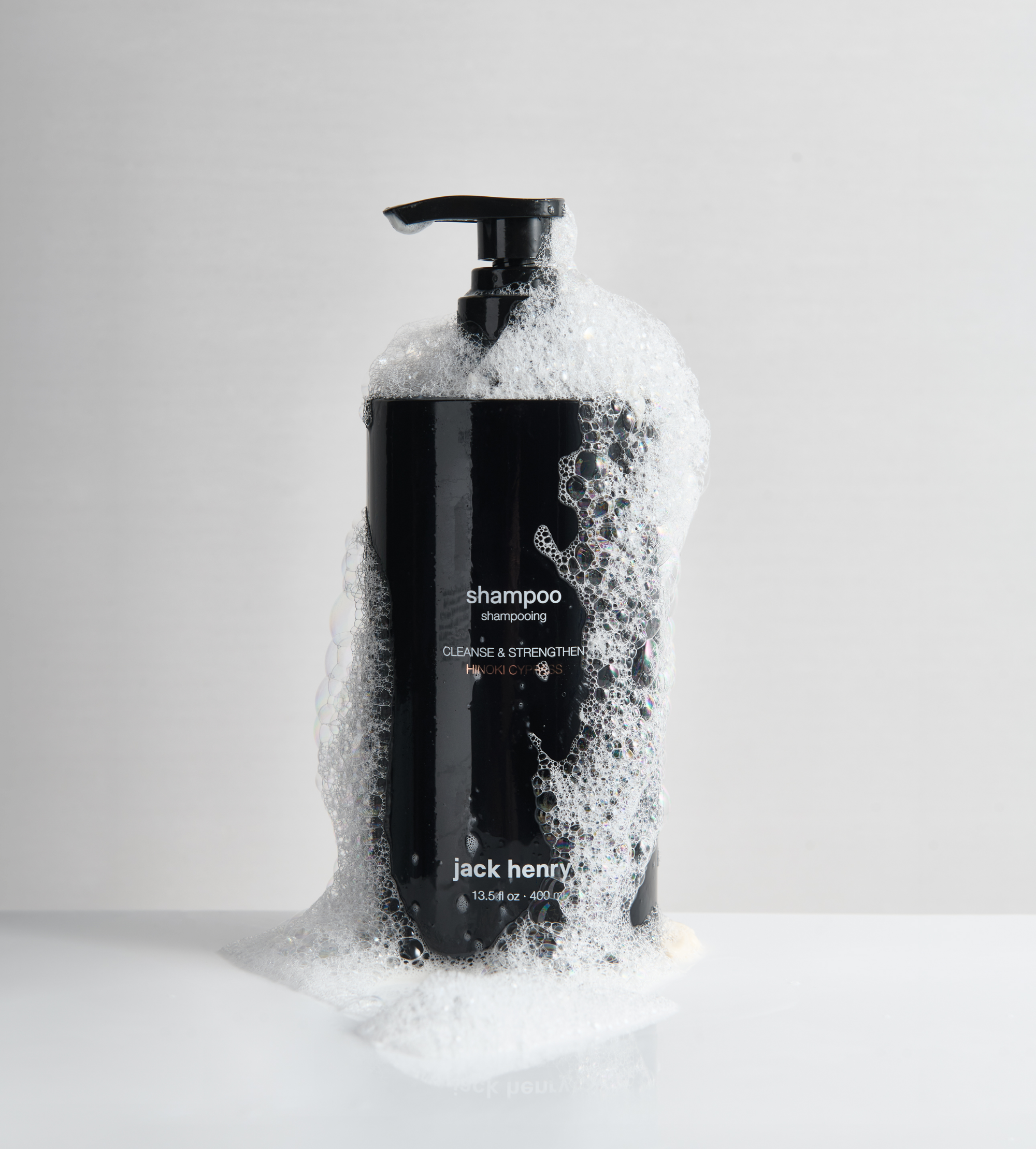 Jack Henry shampoo bottle overflows with cleansing bubbles on a glossy white surface.
