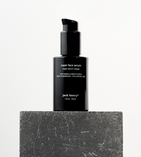 Jack Henry's black bottle of anti-aging Super Face Serum sits on a textured gray stone block.
