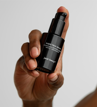 A hand holds a black bottle of Jack Henry Super Face Serum.
