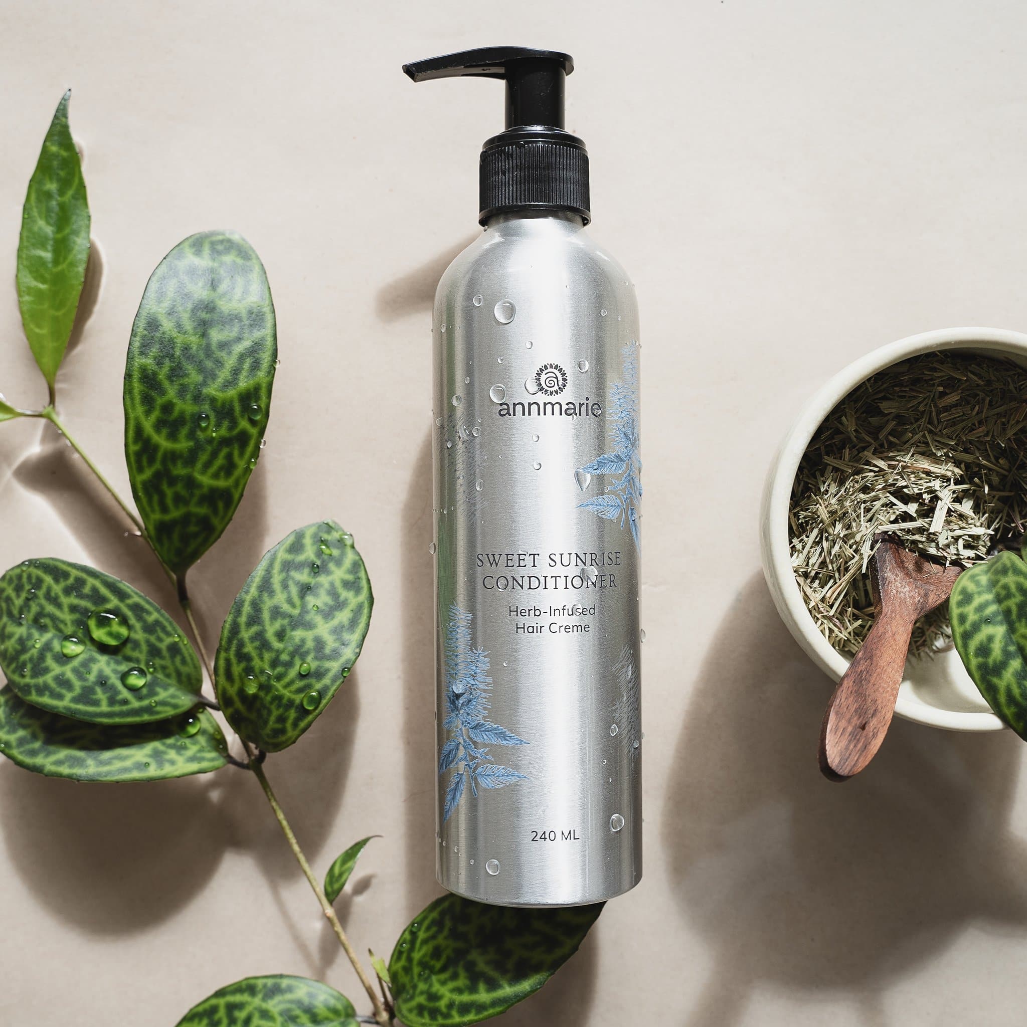 Annmarie's Sweet Sunrise Conditioner bottle sits alongside fresh herbs and a leafy green plant, showcasing natural hair care.
