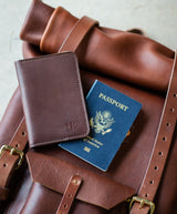 A brown leather travel backpack displays a passport and a matching passport wallet, suggesting international travel.
