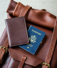 A brown leather travel backpack displays a passport and a matching passport wallet, suggesting international travel.
