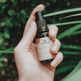A hand holds Annmarie Probiotic Serum, a bio facial elixir, against a green, leafy background.

