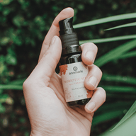 A hand holds Annmarie Probiotic Serum, a bio facial elixir, against a green, leafy background.
