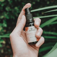 A hand holds Annmarie Probiotic Serum, a bio facial elixir, against a green, leafy background.
