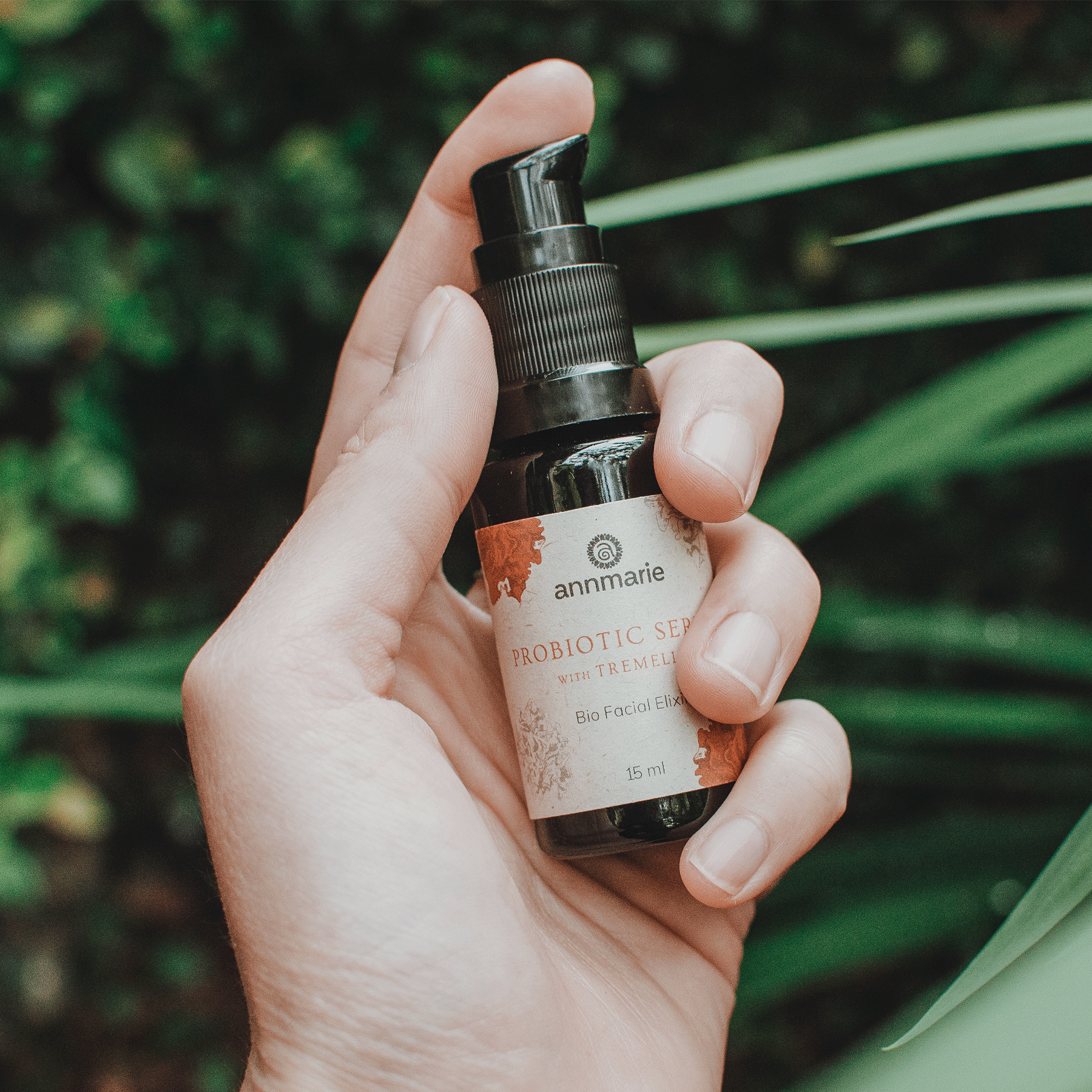 A hand holds Annmarie Probiotic Serum, a bio facial elixir, against a green, leafy background.
