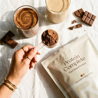 Protein shake ingredients including a glass of chocolate shake, protein powder, and a package of Protein Complete plant-based protein on a white surface.