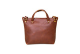 A brown leather tote bag with brass hardware stands isolated on a white background.
