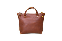 A brown leather tote bag with brass hardware stands isolated on a white background.
