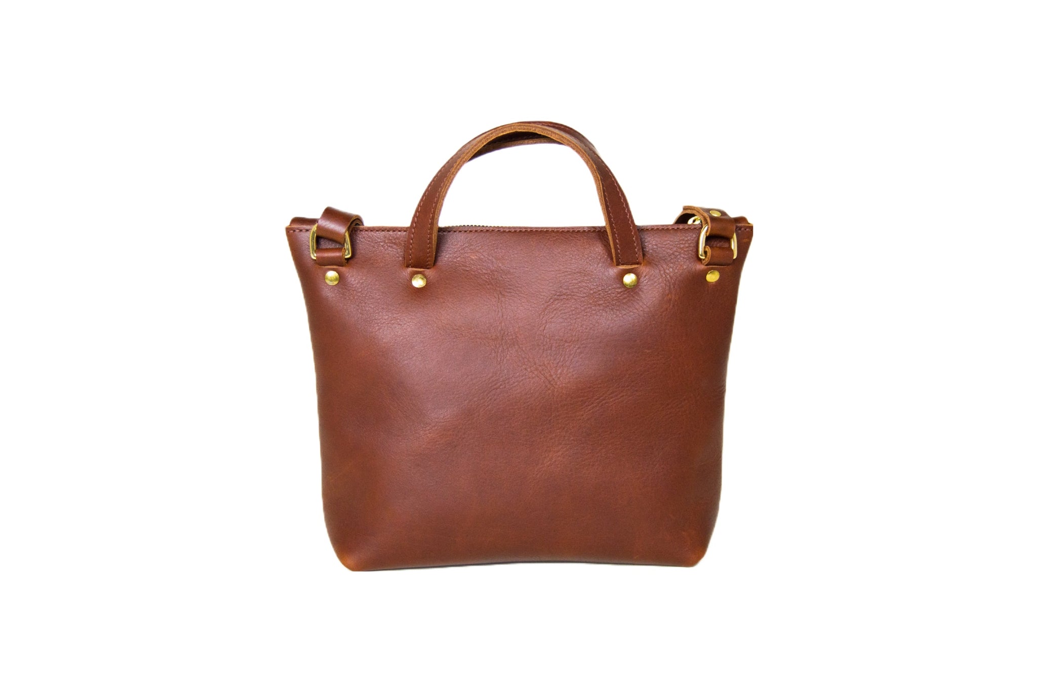 A brown leather tote bag with brass hardware stands isolated on a white background.
