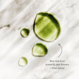 See vibrant green skincare serum droplets, tinted with butterfly pea flower and blue tansy, glistening on a white marble surface.
