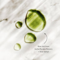 See vibrant green skincare serum droplets, tinted with butterfly pea flower and blue tansy, glistening on a white marble surface.
