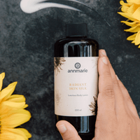 Hand holds Annmarie Radiant Skin Silk luxurious body lotion amid sunflowers on a textured dark surface.
