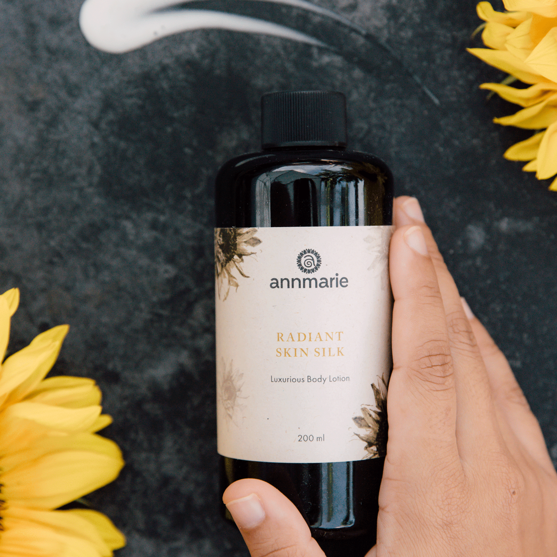 Hand holds Annmarie Radiant Skin Silk luxurious body lotion amid sunflowers on a textured dark surface.

