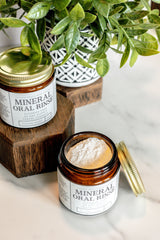 Two jars of Clean Mint mineral oral rinse, a fluoride-free mouth rinse powder, sit next to a leafy green plant on a marble surface.