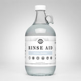 A glass jug displays Rustic Strength Rinse Aid, advertising its hypoallergenic and biodegradable formula for sparkling clean dishes.

