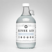 A glass jug displays Rustic Strength Rinse Aid, advertising its hypoallergenic and biodegradable formula for sparkling clean dishes.
