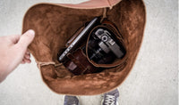 Person opens a brown leather bag revealing a laptop, camera, and leather pouch inside.
