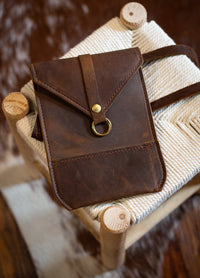 A rustic brown leather crossbody bag rests on a woven rope stool.
