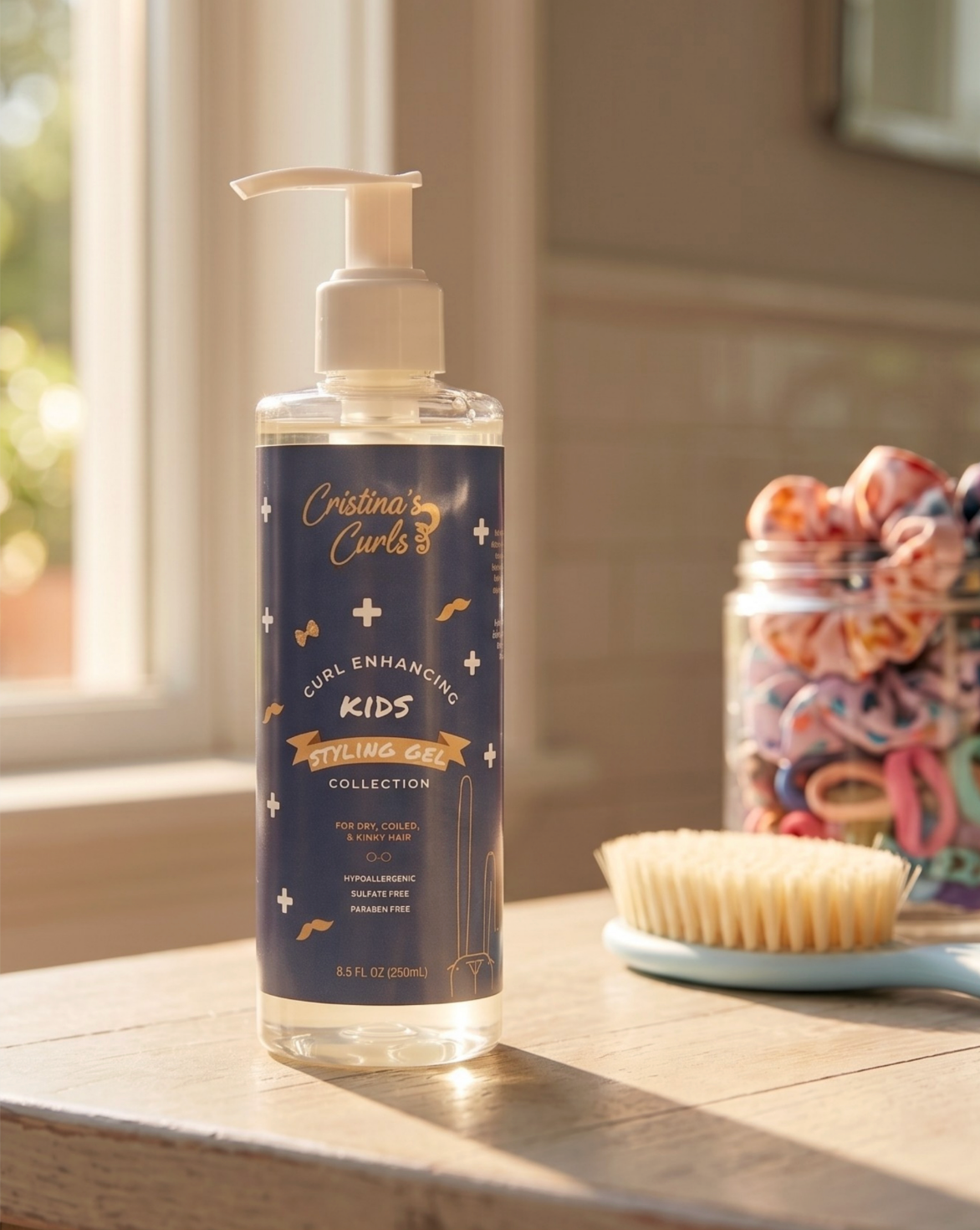A bottle of Cristina's Curls Kids Curl Enhancing Hair Oil Collection stands on a wooden windowsill with warm natural light. The bottle has a dark navy blue label with gold and white text and illustrations.
