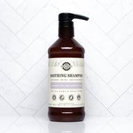 A brown bottle of Rustic Strength Soothing Shampoo with lavender and tea tree essential oils sits on a white tile surface.
