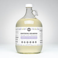 A one-gallon glass jug contains Rustic Strength Soothing Shampoo with lavender and tea tree essential oils, marketed as gentle, non-toxic, and suitable for all ages and hair types.

