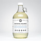A glass jug displays Rustic Strength Soothing Shampoo, highlighting its unscented, hypoallergenic, and plant-derived formula.